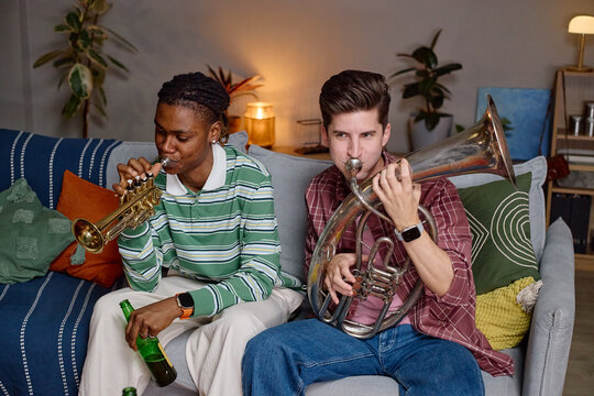 Diverse young adult male football fans playing trumpet and euphonium on sofa while celebrating victory. Image ideal for sports entertainment and fan culture marketing campaigns