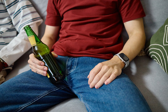 Cropped shot of male football fan sitting on sofa and holding beer bottle. Image represents leisure and friendship, ideal for beverage advertising