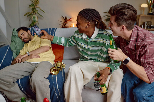 Diverse young adult male football fans laughing and taking photo of sleeping friend on sofa. Image represents friendship and humor, ideal for social media marketing campaigns