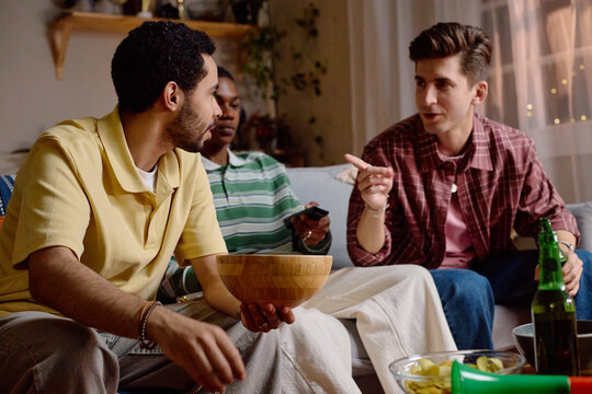 Diverse young adult male football fans sitting on sofa, talking and watching game on television. Image suitable for sports broadcasting, entertainment, and social gathering campaigns