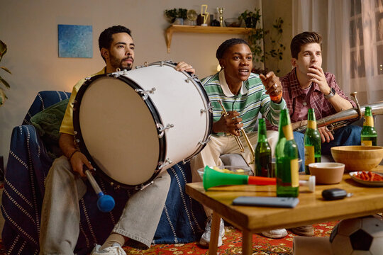 Diverse young adult male football fans watching match at home with musical instruments. Image ideal for sports broadcasting promotions and entertainment marketing campaigns