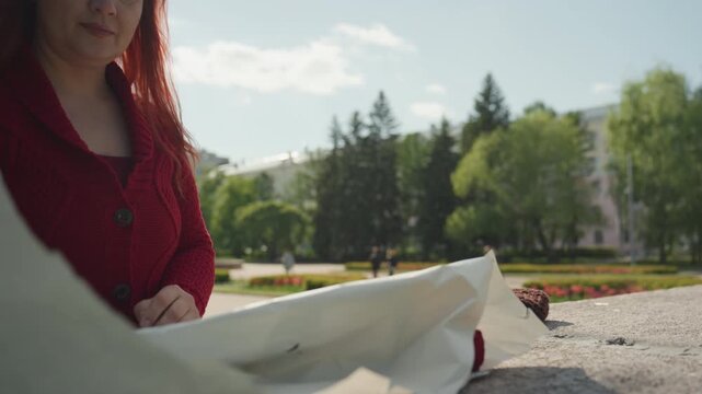 Close up of woman with long red hair wearing textured knit sweater lying on pavement and interacting with white paper. Woman arranges rose stem alongside other objects in outdoor park