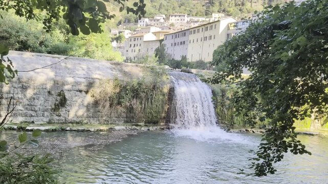 Cascata naturale che si riversa in uno specchio d'acqua limpida, ai piedi di un caratteristico borgo storico italiano immerso nel verde della natura.
