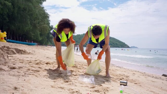 Two volunteers collecting plastic bottles and trash on a sandy beach. 