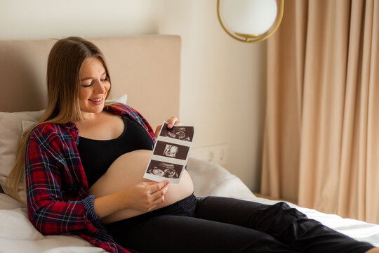 Pregnant woman smiling on bed while looking at ultrasound photos and touching her baby bump