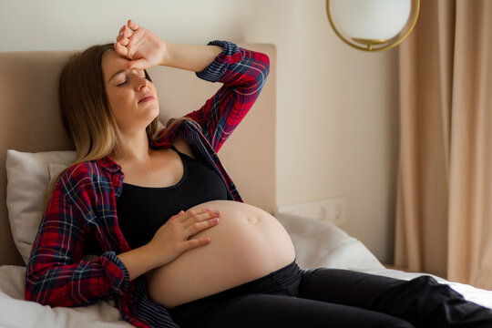 Pregnant woman resting on bed with hand on belly feeling tired during late pregnancy at home