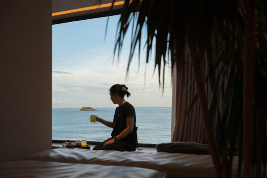 The woman is sitting by the window, holding a drink.