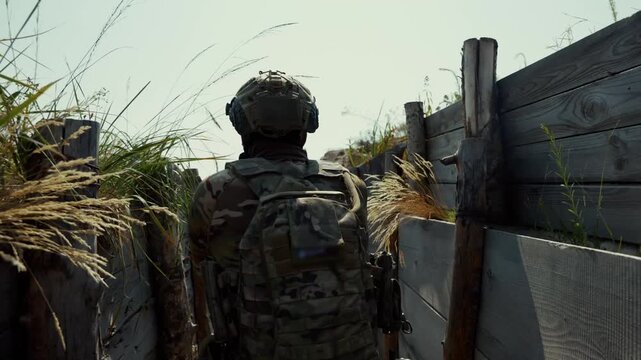 Back view of armed soldier in full camouflage tactical gear and helmet taking firing position at edge of trench fortification among dry grass in combat zone. Military warfare concept for defense, conf