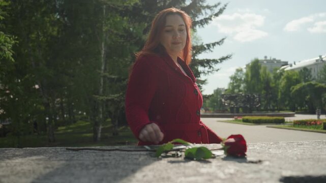 Woman in red coat approaches and interacts with objects on outdoor stone surface placing or adjusting items including leaves. scene features soft natural light against green trees distant