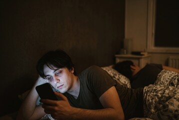 Obraz premium A man is lying in bed at night. He holds his phone and stares at the screen. Another person is lying beside him in the same bed, not looking at the phone