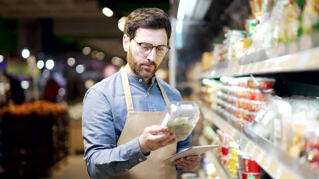 Male supermarket worker using digital tablet checking shelves with products in store. Mature manager in apron inspects goods, writes an inventory list, and controls stock availability in retail market