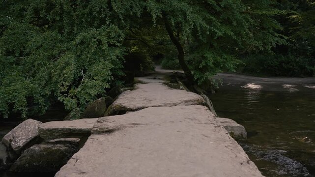 Tarr Steps showing the iconic clapper bridge stretching across the River Barle. The scene emphasises stillness and natural beauty, subtle movement in the water and leaves enhancing the peaceful scene.