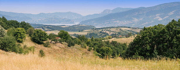 Naklejka premium Panoramic hillside view sweeping across layered green valleys, golden meadow, scattered farmhouses, and a distant Danube framed by misty mountains. Travel concept.