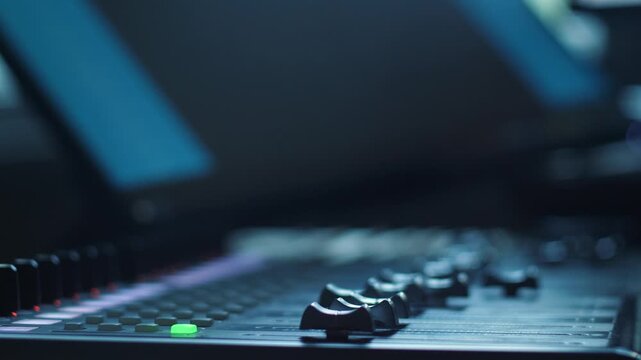Close-up of a professional sound mixing console in a recording studio with a green illuminated button and blurred background screens. 