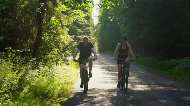 Active couple cycling together along a peaceful sunlit forest road, enjoying outdoor exercise, summer fitness, and nature adventure in lush green woodland scenery