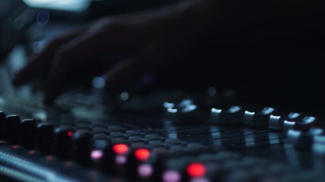 Close-up of a sound engineer's hand adjusting faders and controls on a professional mixing console. The process of mixing and recording audio in a dim studio with illuminated control buttons.