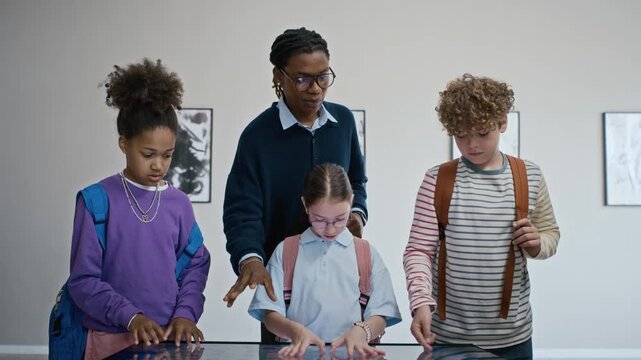 Black male teacher guiding diverse school kids using interactive digital touch screen table in art gallery. Video perfect for educational technology and museum field trip marketing