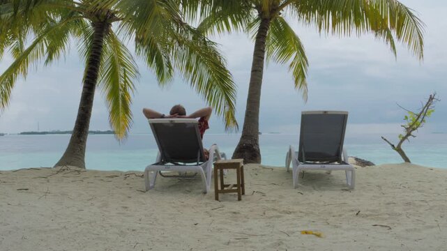 A vacationer rests with his hands behind his head on a sun lounger beside a second empty lounger, facing a calm turquoise lagoon from a palm-shaded Maldives beach in a static wide shot.