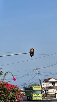 Vertical static of a flashing yellow amber traffic light on a pole with passing traffic and clear blue sky background.
