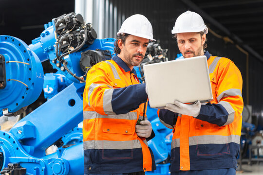 Robotics engineers using laptop to program industrial robot arm in factory, Automation technicians working together with computer on robotic machinery, Engineers inspecting automated robotic system