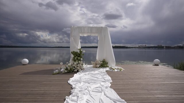 Lakeside wedding arch with flowing white aisle runner on wooden dock beneath storm clouds, framed by floral decor, reflective water, calm shoreline, and elegant outdoor ceremony setting