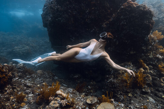 Woman freediving in white swimsuit above a coral reef in blue sea.
