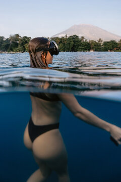 Woman swims in tropical sea with underwater view and volcanic mountain landscape in the background