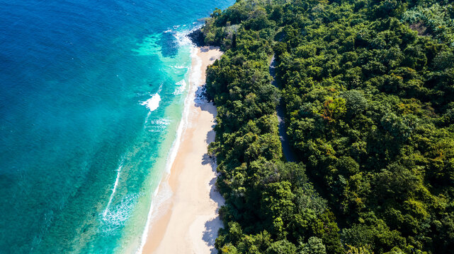Aerial view of a pristine sandy beach meeting turquoise waters, bordered by lush tropical forest and a winding coastal road in Bajawa, Nusa Tenggara Timur, Indonesia.
