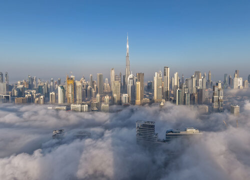 Aerial view of Burj Khalifa rising above a thick layer of morning fog covering the modern skyscrapers and urban skyline of the city Dubai, Dubai, United Arab Emirates.