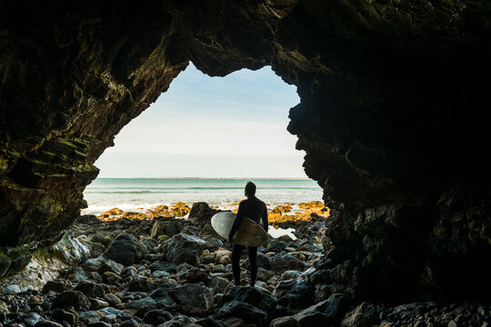 France, Bretagne, Finistere, Crozon peninsula, man standing on rocky beach with surfboard