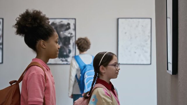 African American girl and Caucasian girl looking at modern paintings in art gallery during school field trip, pointing at artwork. Perfect for promoting museum youth programs and art education