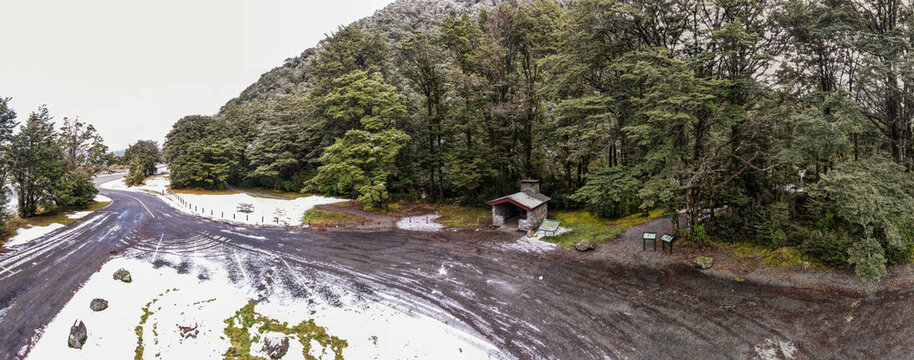 Aerial view of a snow-covered roadside parking area and a small stone shelter nestled within a dense forest in the Canterbury Region, New Zealand.