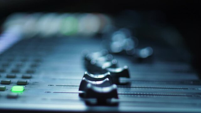 Macro shot of automated faders and knobs moving on a professional digital audio mixing console. The mixing board controls adjust volume levels dynamically during music post production 
