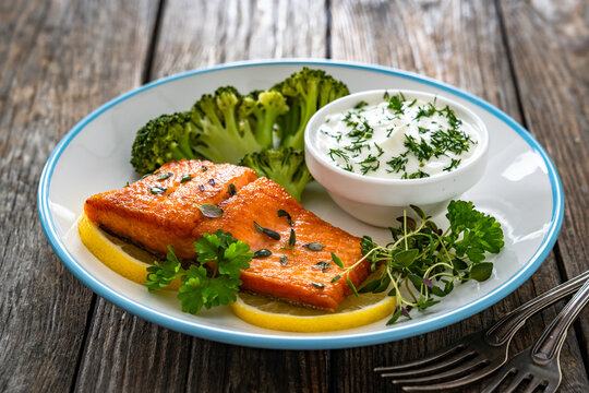 Fried salmon steak with boiled broccoli and dill yogurt sauce on wooden table
