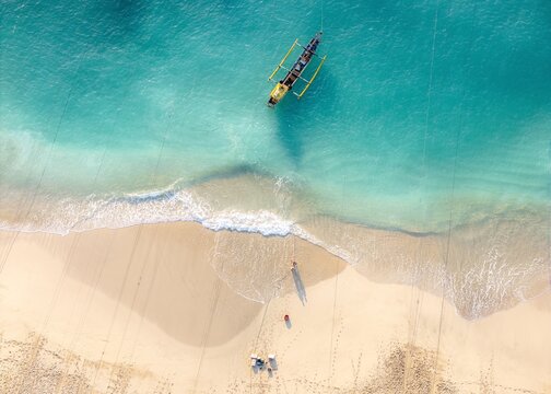 Aerial view of a traditional outrigger boat floating in turquoise water near the sandy shore of Papuma Beach with people walking on the sand in Jember, East Java, Indonesia.