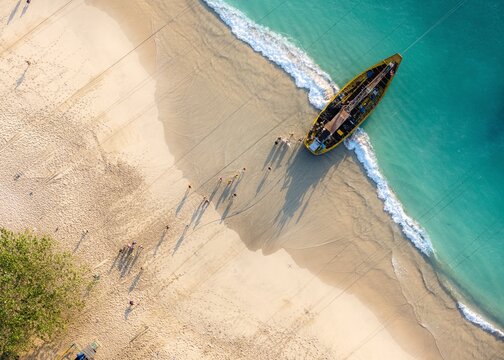 Aerial view of Papuma Beach with a yellow fishing boat resting on the sandy shore, turquoise waves lapping the coast, and people walking along the beach in Jember, East Java, Indonesia.