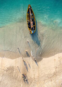 Aerial view of a yellow fishing boat resting on the sandy shore of Papuma Beach with people walking nearby in the clear turquoise water in Jember, East Java, Indonesia.
