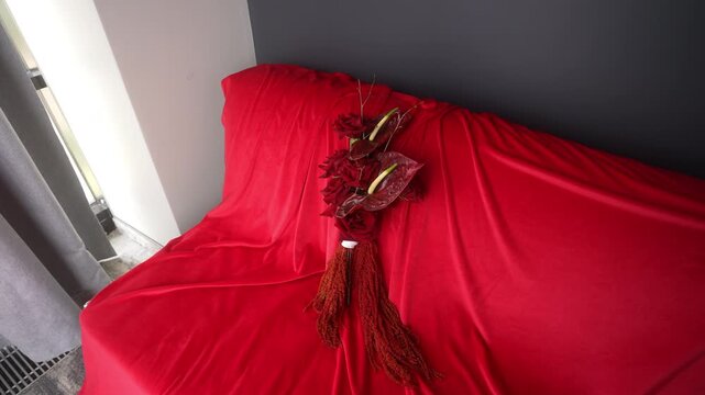 Red draped sofa with centered floral bouquet and tassel settling on upholstery under soft daylight, with red, fabric, rose, anthurium detail, showing bouquet, floral, flower, context
