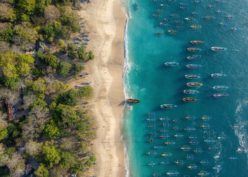 Aerial view of Papuma Beach with numerous traditional fishing boats anchored in the turquoise waters along the sandy coastline and lush green forest in Jember, East Java, Indonesia.
