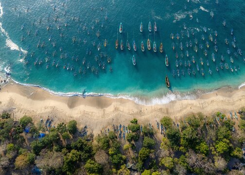 Aerial view of Papuma Beach with numerous traditional fishing boats anchored in turquoise water along the sandy coastline surrounded by lush tropical vegetation in Jember, East Java, Indonesia.