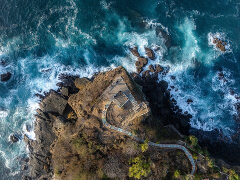 Aerial view of Papuma Beach featuring a rocky cliff with a walking path surrounded by crashing waves and deep blue ocean in Jember, East Java, Indonesia.