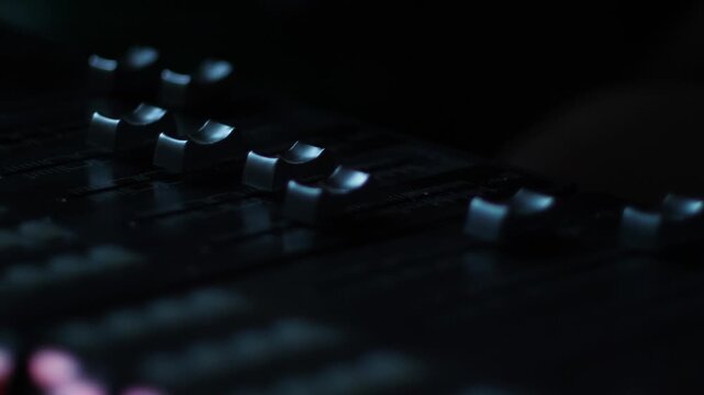 Close-up shot of audio faders on a digital mixing console in a dark recording studio. Automated controls move smoothly during sound mixing and post-production mastering