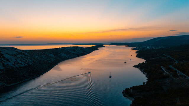 Aerial view of the coastal landscape near Rab at sunset, with a boat leaving a wake in the calm water, surrounded by rocky hills and the Adriatic Sea in Rab, Primorje-Gorski Kotar, Croatia.
