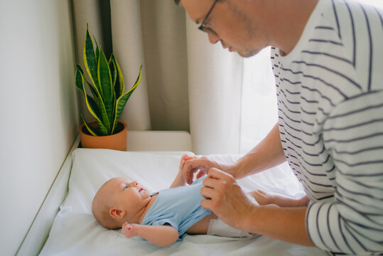 Young father carefully dressing his infant son on a white changing table, putting on a blue bodysuit while providing loving care and bonding during their morning routine