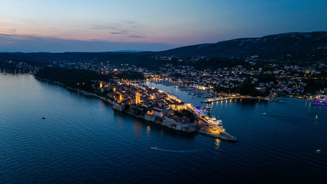 Aerial view of Old Town Rab illuminated at night with historic stone buildings, church towers, and a bustling harbor along the Adriatic Sea coastline, Rab, Primorsko-goranska zupanija, Croatia.