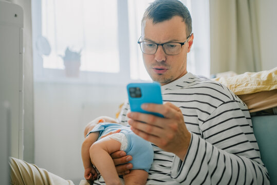 Father holding his infant child while focused on a smartphone, representing the daily challenges and modern realities of working from home and remote work