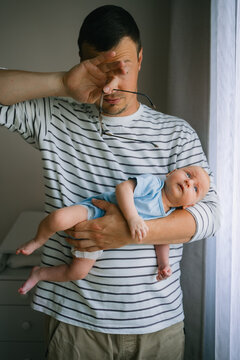Tired father experiencing parental stress and exhaustion, rubbing his eyes while babysitting a newborn baby, struggling with the demands of infant care