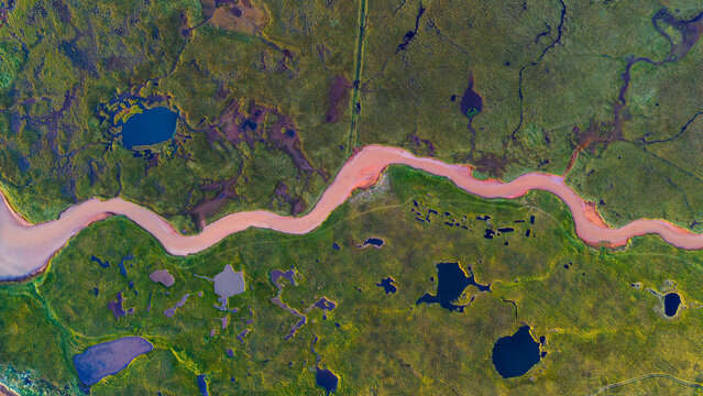 Aerial view of a winding river with reddish water flowing through a vibrant green landscape dotted with small ponds near Selfoss, Sveitarfelagid Arborg, Iceland.