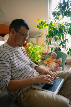 Young father working on a laptop from home, sitting on the floor while his infant son rests in a baby bouncer nearby, navigating parenthood and professional commitments
