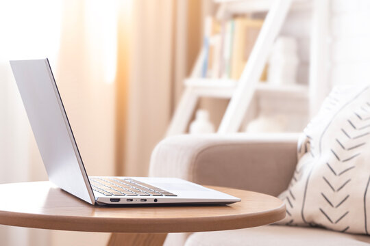 Open modern laptop on wooden round table on blurred interior background of sofa and curtains in living room